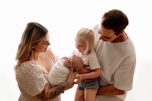 Family of four with mom holding newborn baby, dad carrying toddler girl, posing together on white background.