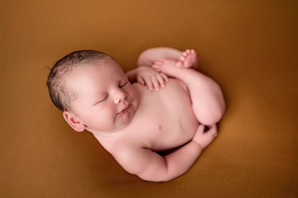 Sleeping newborn baby curled in a relaxed pose on a warm golden-brown blanket during a professional newborn photography session.