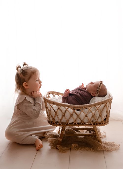 A toddler girl in a beige knit outfit gazes adoringly at a swaddled newborn baby resting in a wicker bassinet.