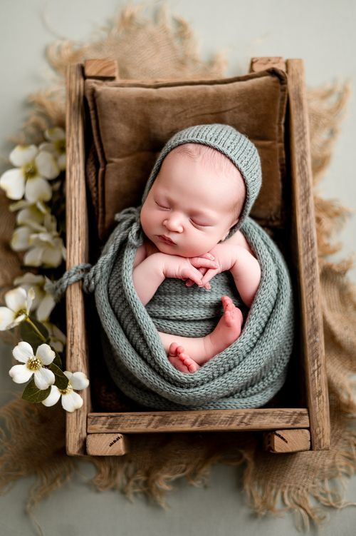 Sleeping newborn wrapped in sage green knit swaddle and bonnet rests in rustic wooden crib prop.