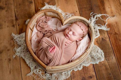 Newborn baby swaddled in pink wrap sleeping in heart-shaped wicker basket on wooden floor with lace.