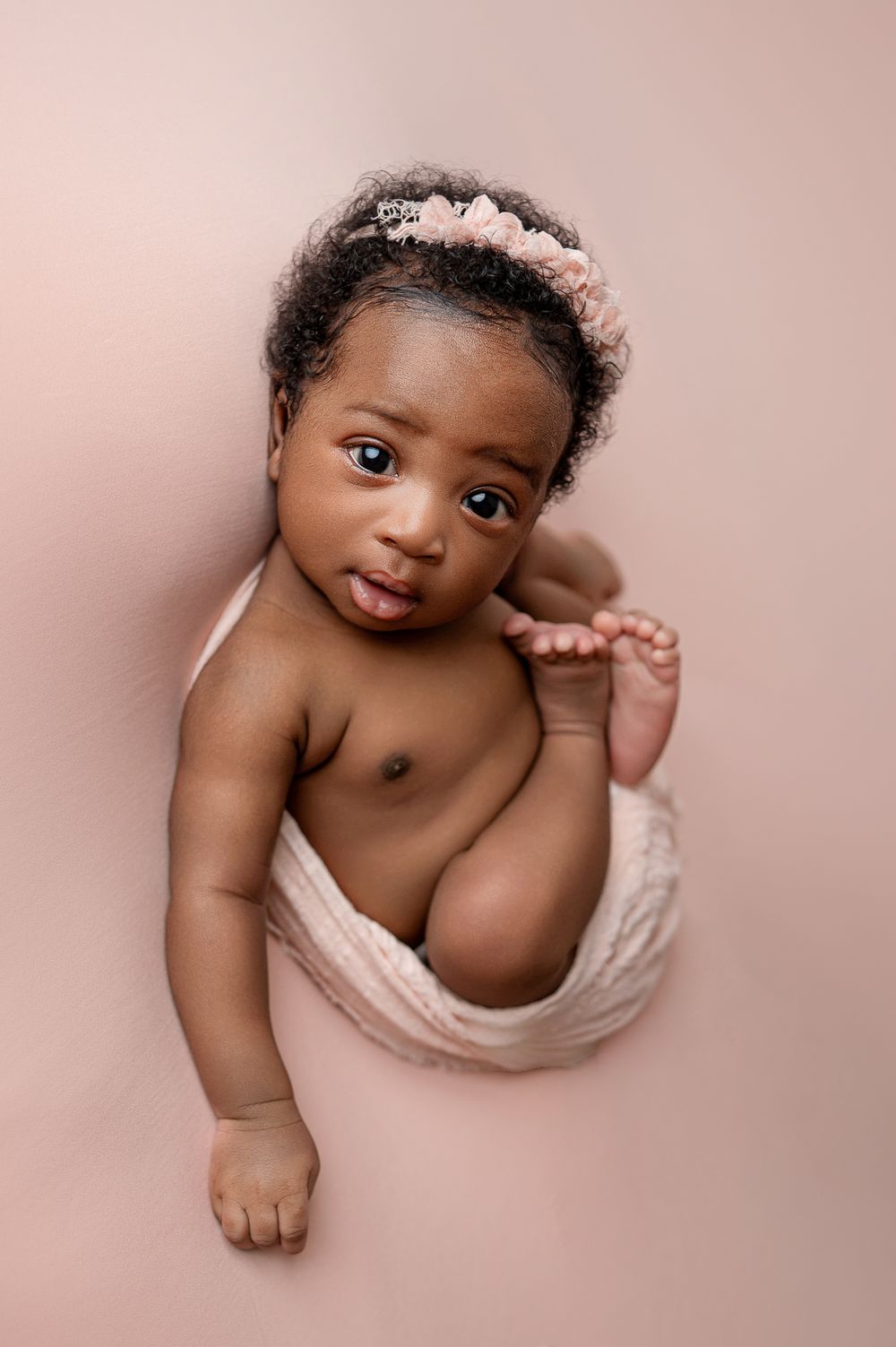 Adorable baby girl wearing a pink floral headband, wrapped in a soft blush wrap, looking up during a newborn photo session.