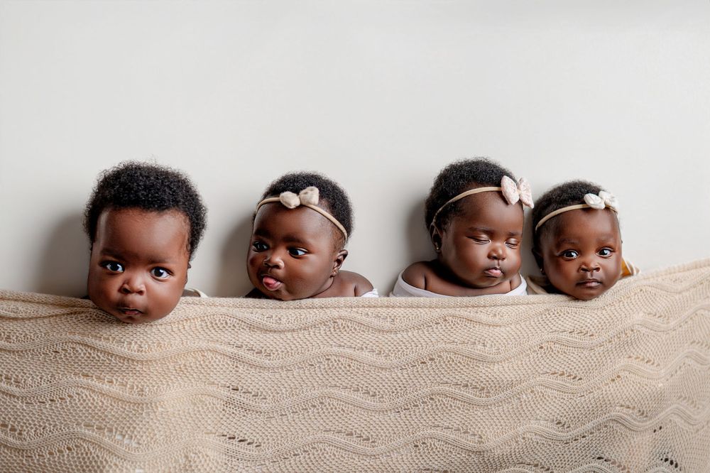 Four adorable Black babies peek over a knit beige blanket, three wearing floral headbands, against a white background.