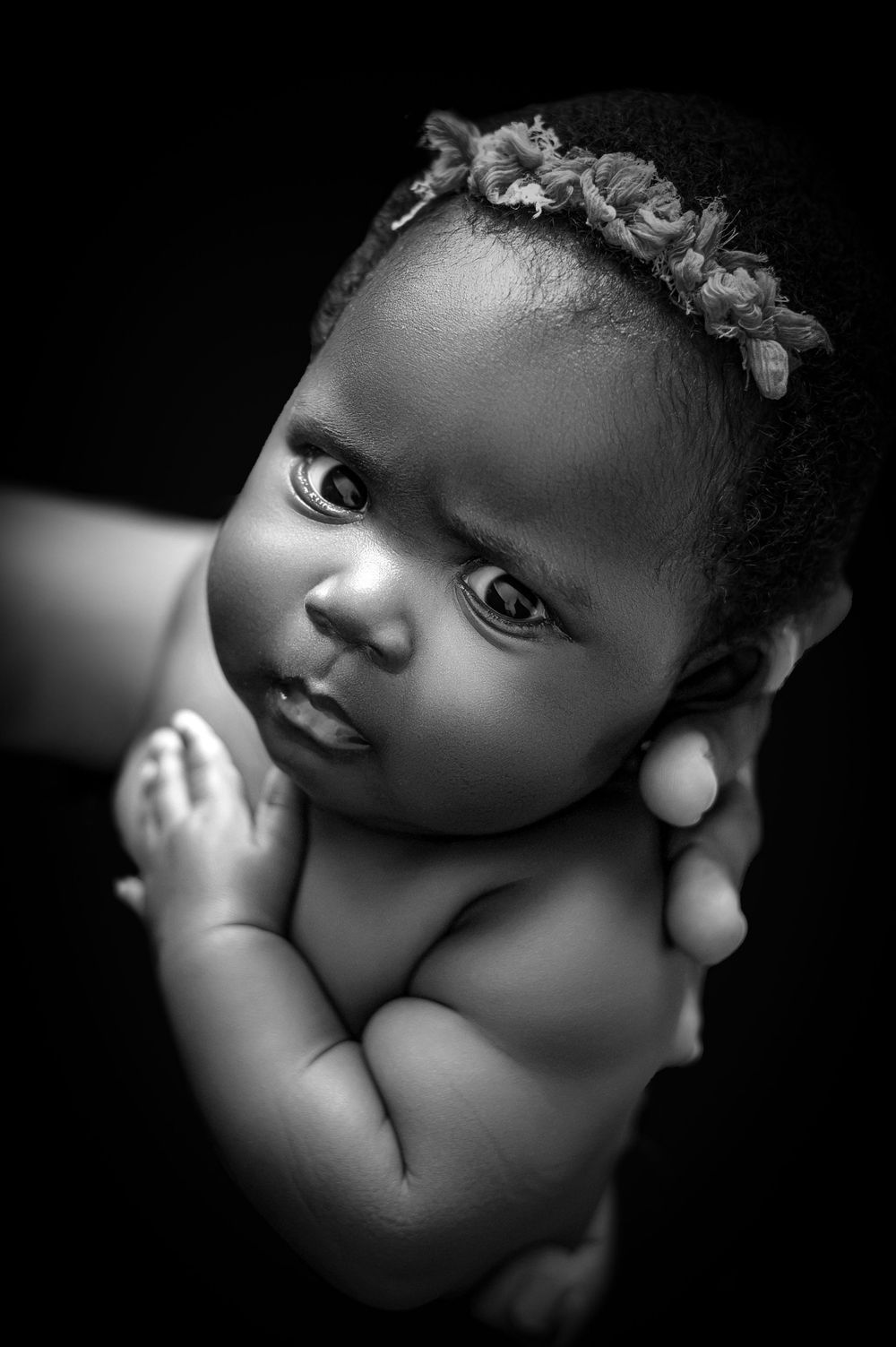 Black and white portrait of a newborn baby girl wearing a floral headband, held gently by adult hands.