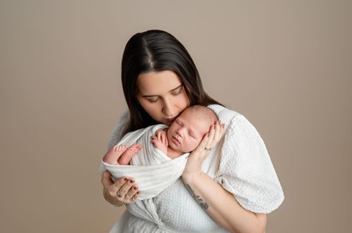 A mother tenderly holds her sleeping newborn baby wrapped in white against her chest in a studio portrait.