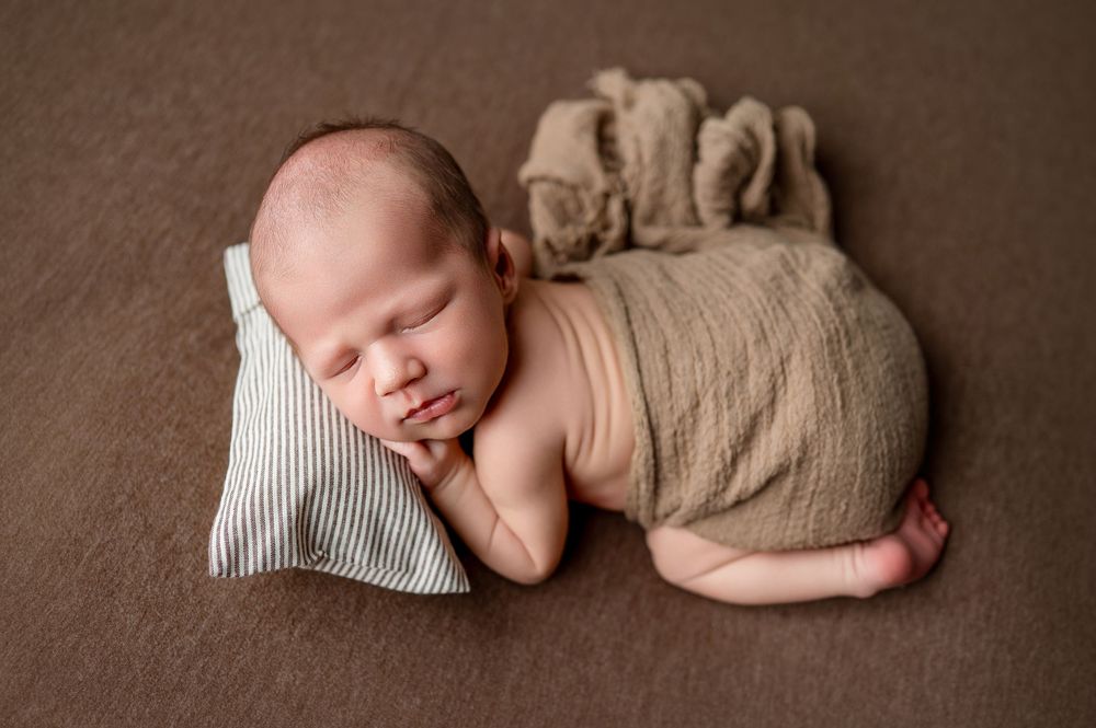 Newborn baby sleeping peacefully wrapped in tan fabric on a brown blanket with a striped pillow.