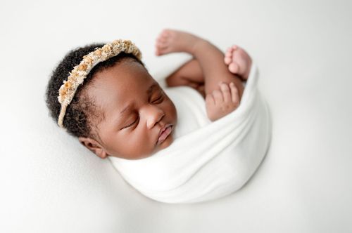 Sleeping newborn baby wrapped in white swaddle with floral headband on white background.
