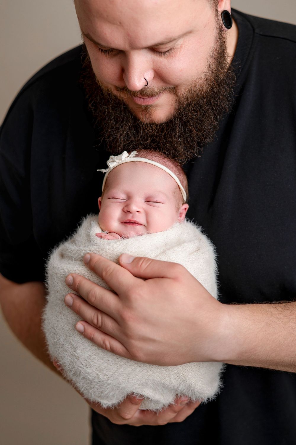 Bearded father in black shirt smiling down at swaddled newborn baby girl wearing white bow headband.