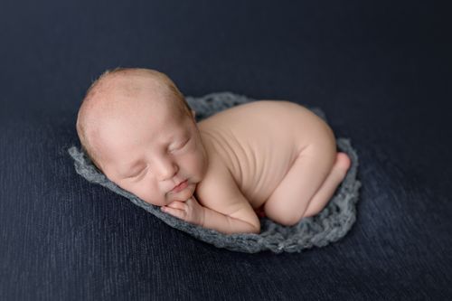 Peaceful newborn baby curled up on gray crochet bowl on dark background.