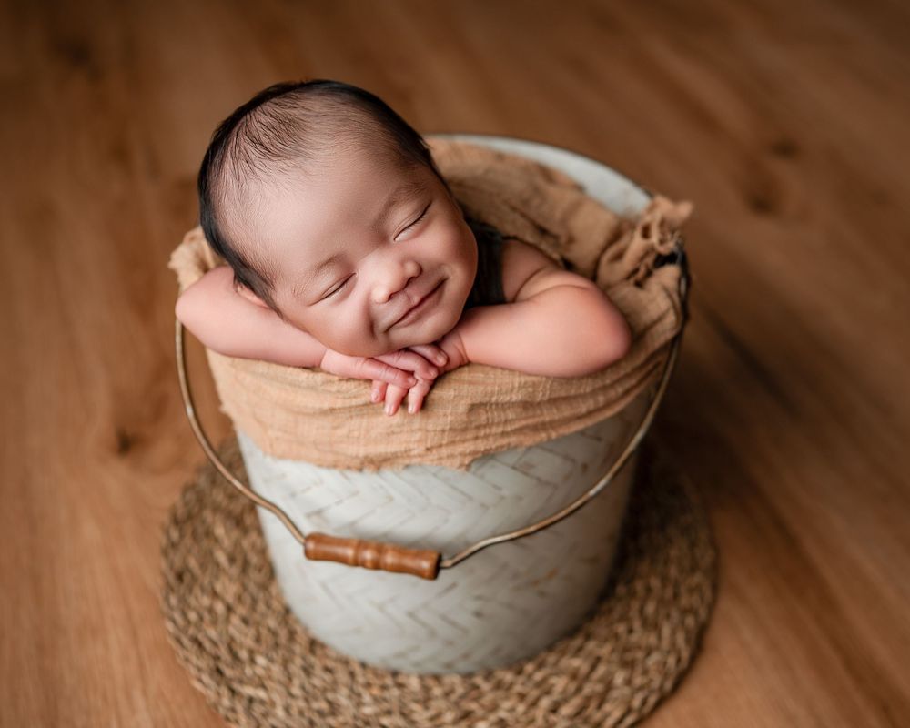 Smiling newborn baby resting chin on hands inside a decorative white bucket on a wooden floor.