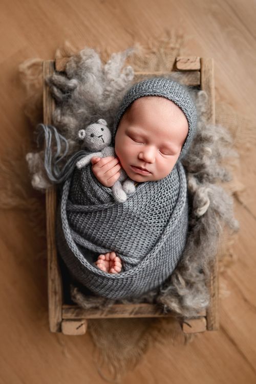 Newborn baby swaddled in gray knit wrap holding small teddy bear in rustic wooden crate.