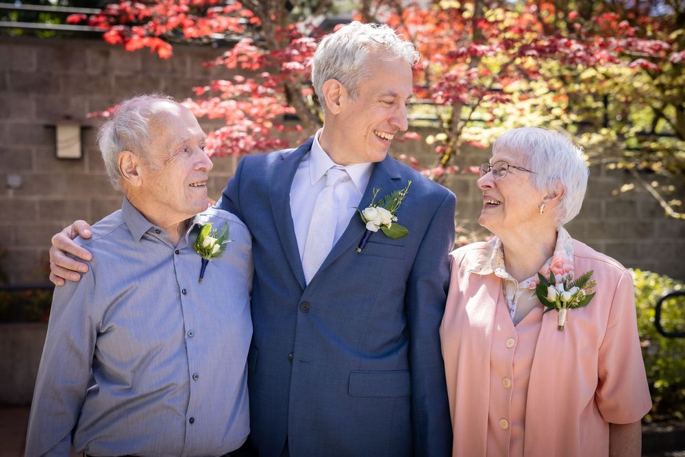 Groom in blue suit poses with elderly parents wearing boutonnieres outdoors near red autumn foliage at wedding.
