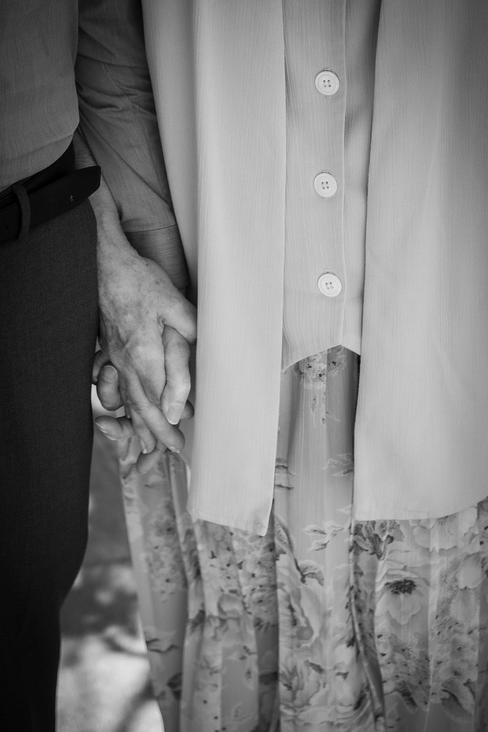 Black and white photo of elderly couple holding hands, woman in floral skirt and button-up blouse, man in dark trousers.