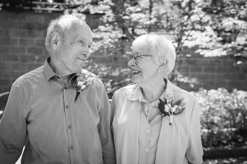Elderly couple smiling at each other outdoors, wearing boutonnieres, in a black and white photo.