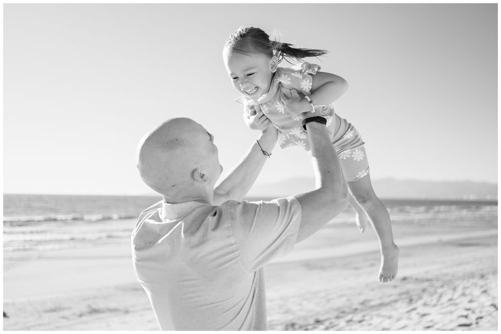 Man lifting laughing child high in the air on a sunny beach, black and white family photo in Punta Mita.