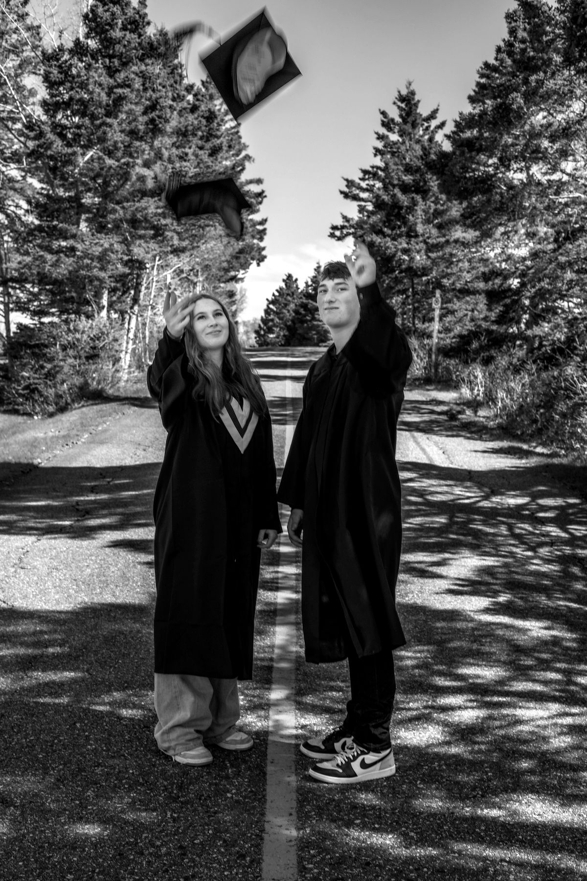 Two graduates in black robes toss their caps on a tree-lined road in a black and white photo.