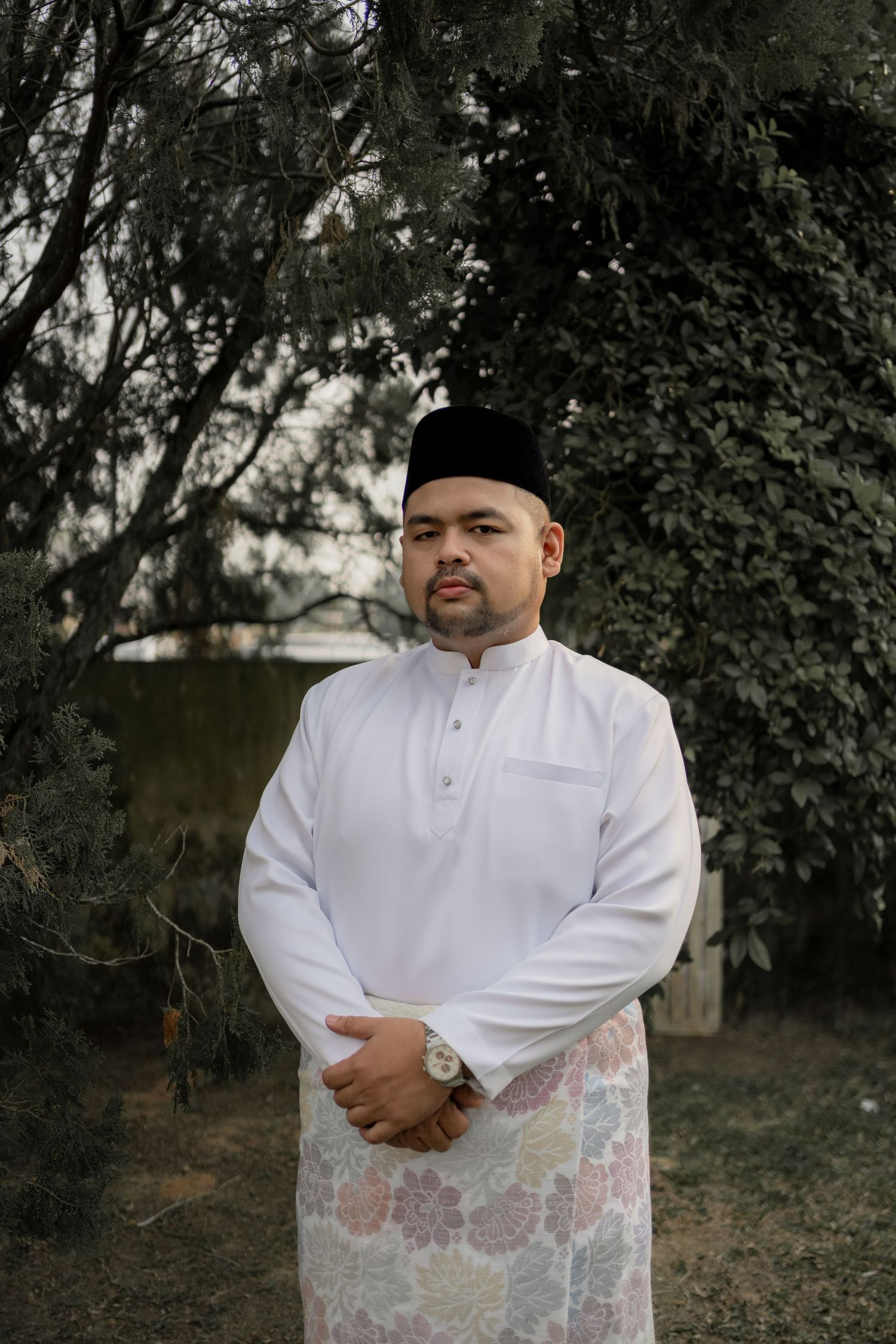 A person in traditional Malay attire stands against a backdrop of green foliage.
