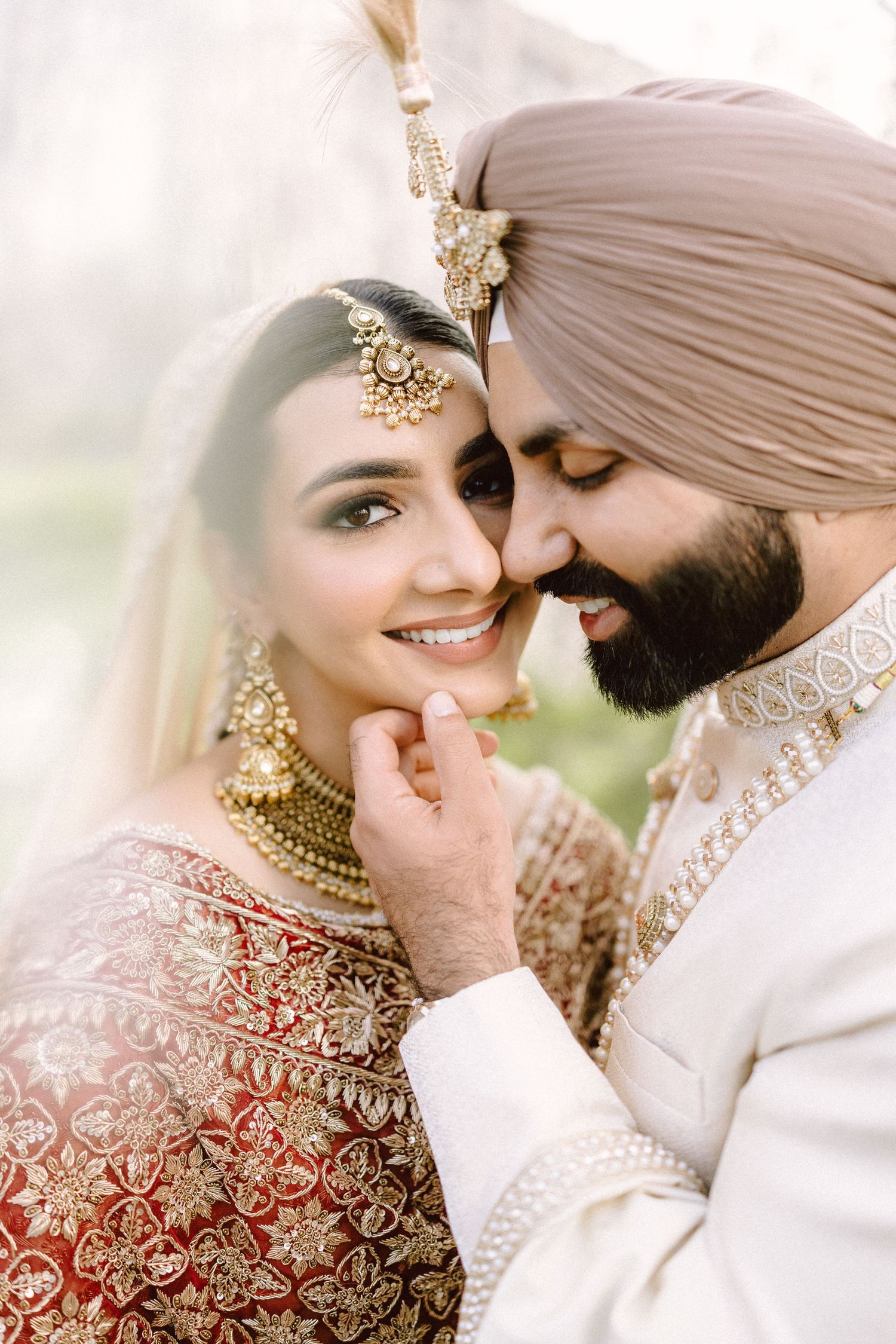 A Sikh bride and groom in traditional wedding attire share a tender moment, adorned with gold jewelry and a turban.