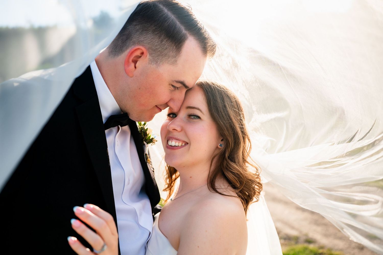 A bride and groom share a romantic moment as her veil billows in the wind during their outdoor wedding photo session.