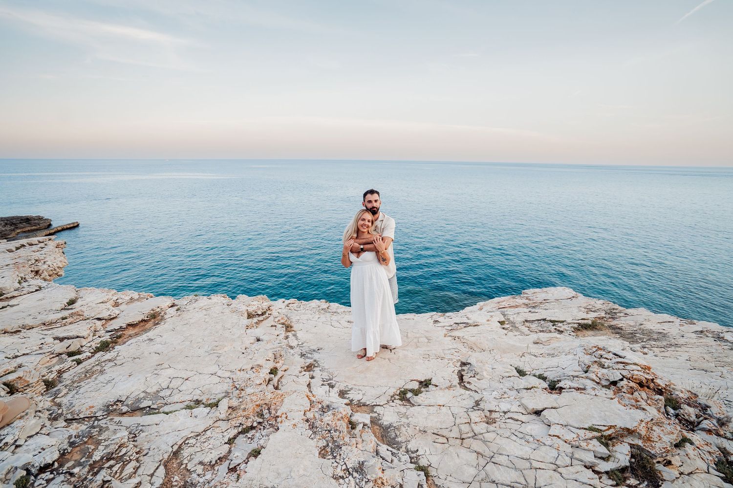 Person in white dress standing on rocky coastal cliff with turquoise sea and pastel sky in background.