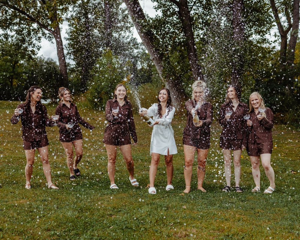 Bride in white romper celebrates with six bridesmaids in maroon rompers, tossing confetti outdoors on green grass.