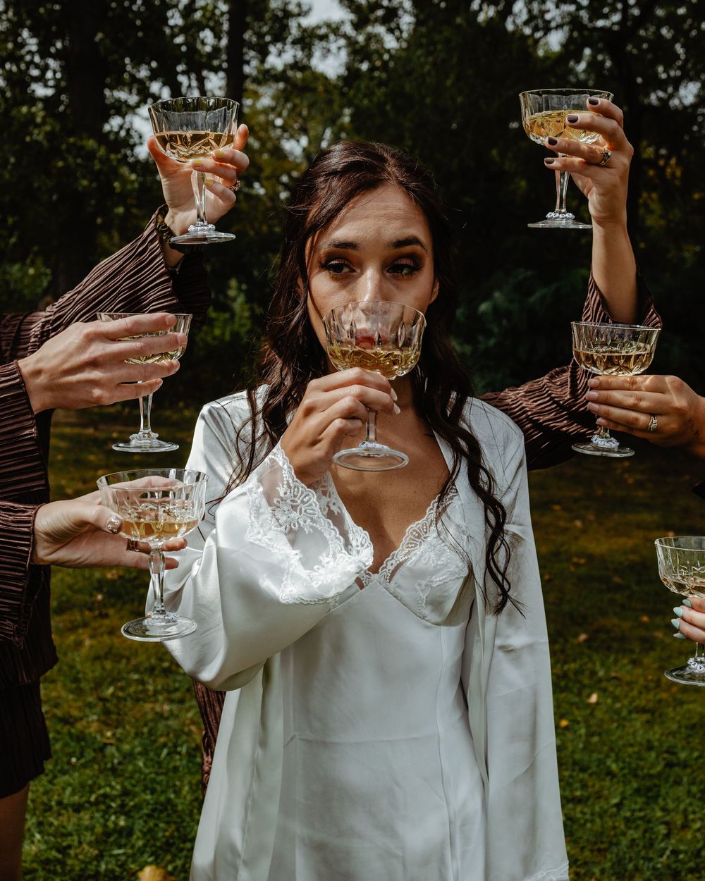 Bride in white robe toasting champagne coupes with bridesmaids outdoors in a garden setting.