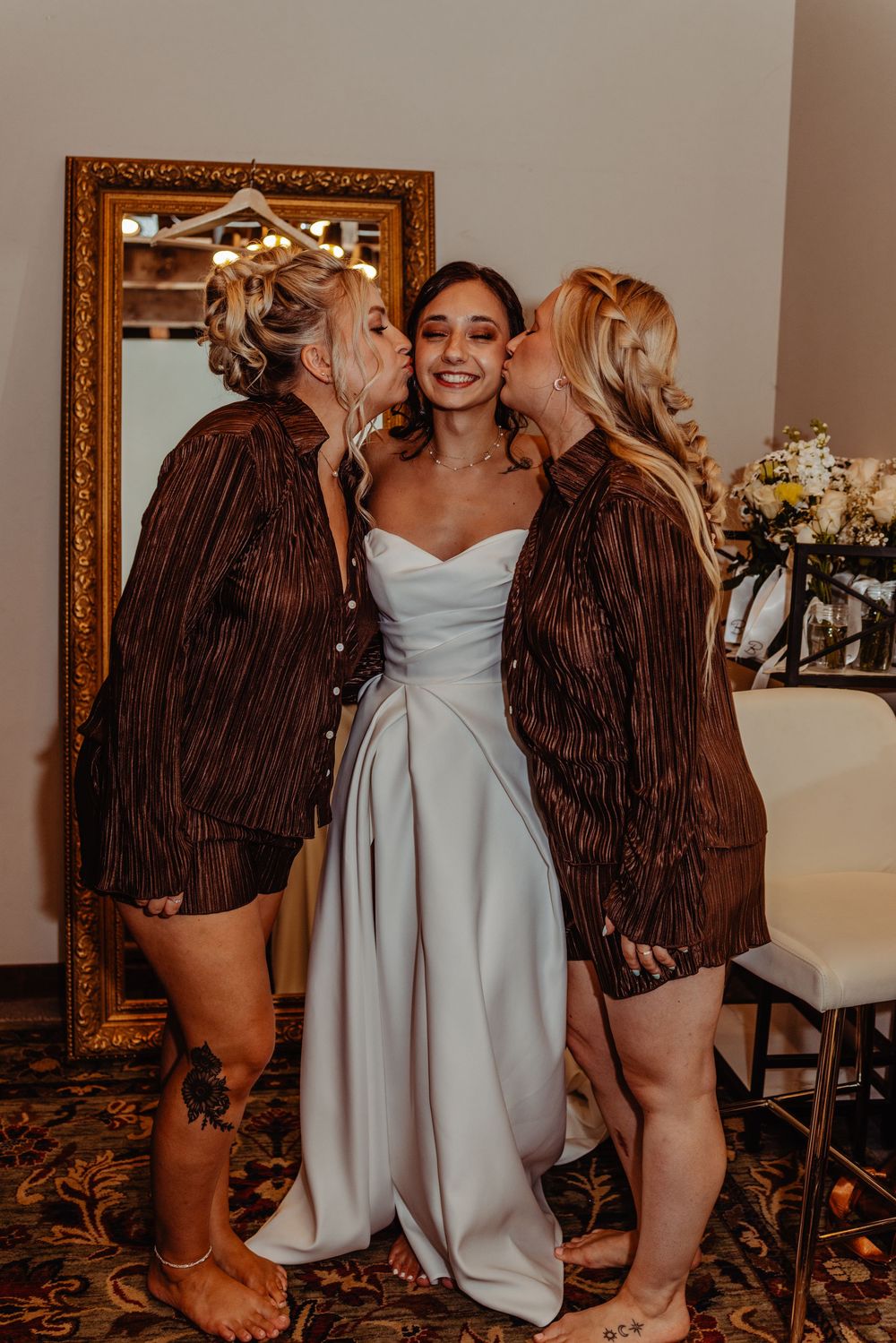 Bride in white gown smiles as two women in brown jackets kiss her cheeks near a gold mirror.