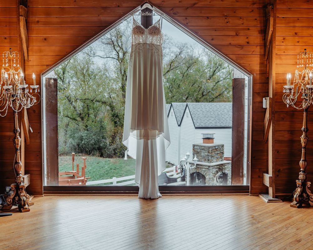 A wedding dress hanging in a rustic barn window flanked by elegant crystal candelabras with a scenic outdoor view.