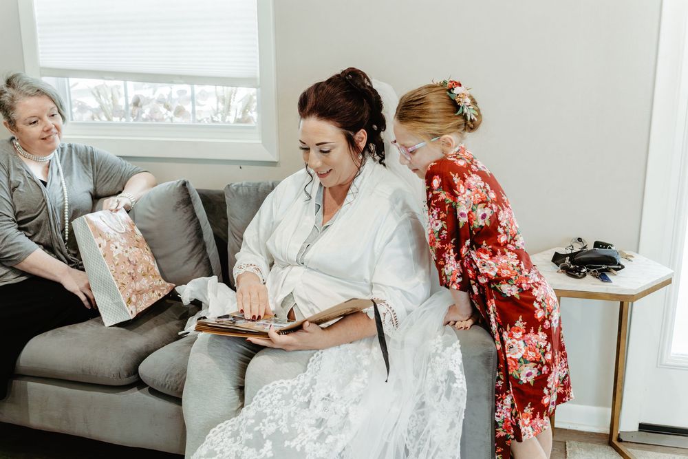 Bride in white robe reviews wedding album with bridesmaids on gray sofa during bridal preparations.