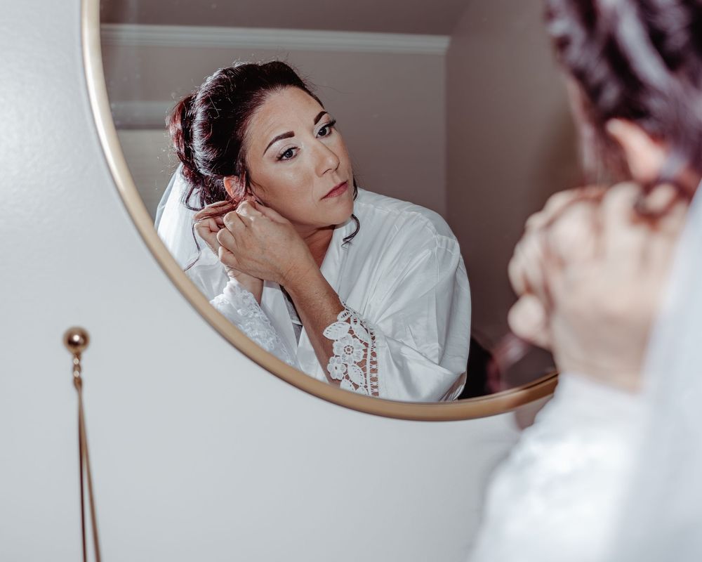 Bride in white lace robe puts on earrings while checking her reflection in round gold mirror.