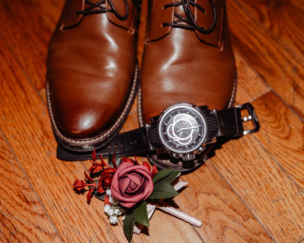 Brown leather dress shoes, black chronograph watch, and burgundy floral boutonniere arranged on hardwood floor.