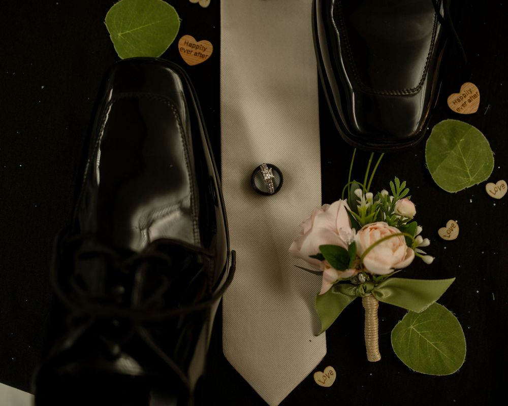 Black dress shoes, white tie, ring, and boutonniere with pink roses arranged on dark background for wedding.