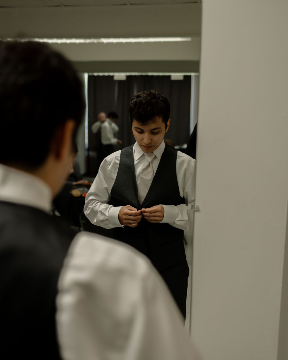 Young boy in white shirt and black vest buttoning up while looking down, reflected in a mirror.