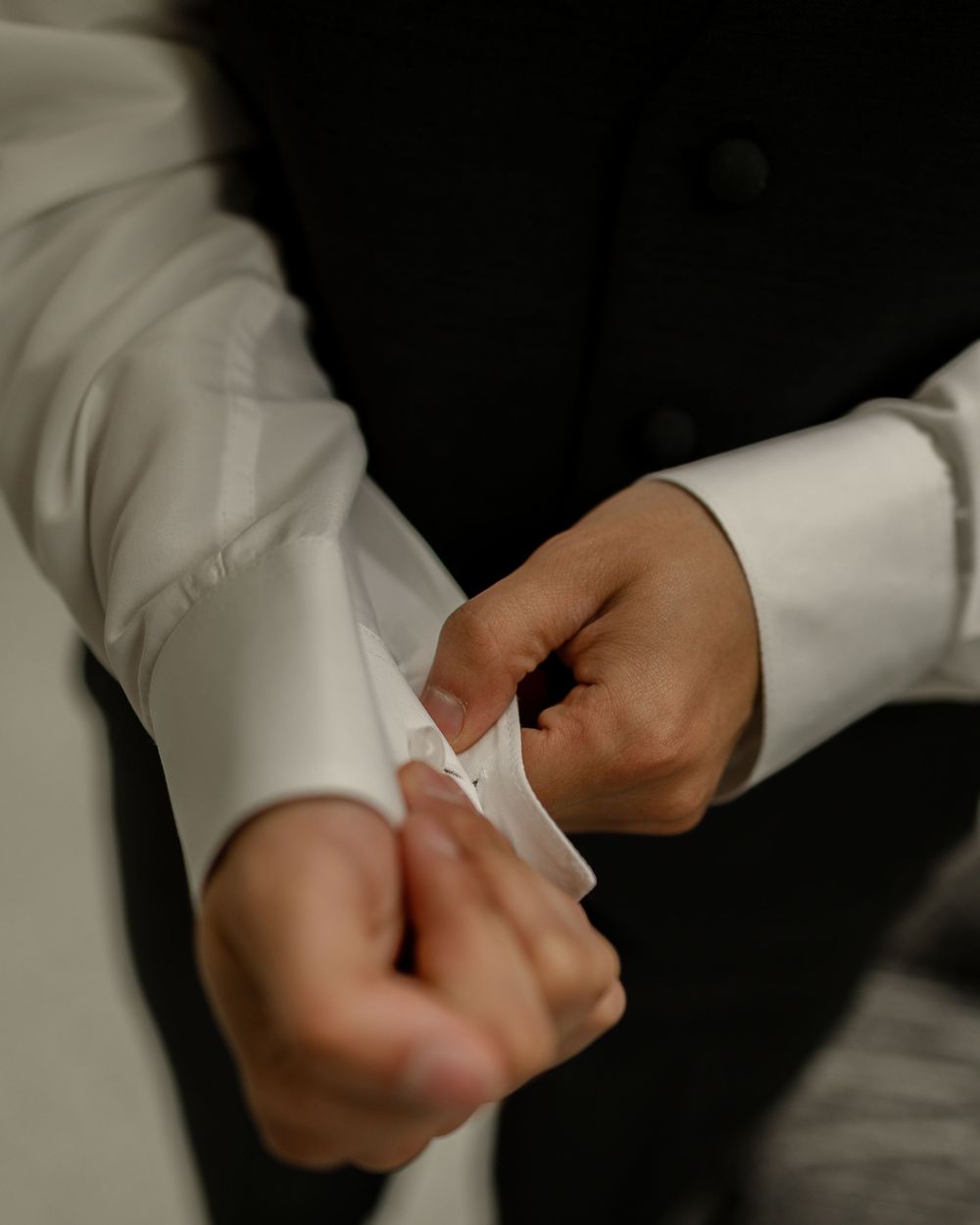 A man in a white dress shirt and black suit fastening his cuff links, preparing for a formal event.