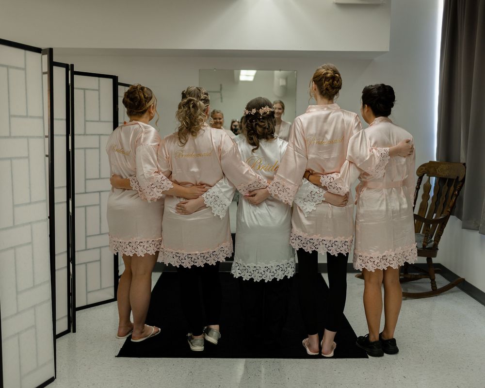 Bridesmaids in matching blush satin robes pose together facing a large mirror during wedding preparations.
