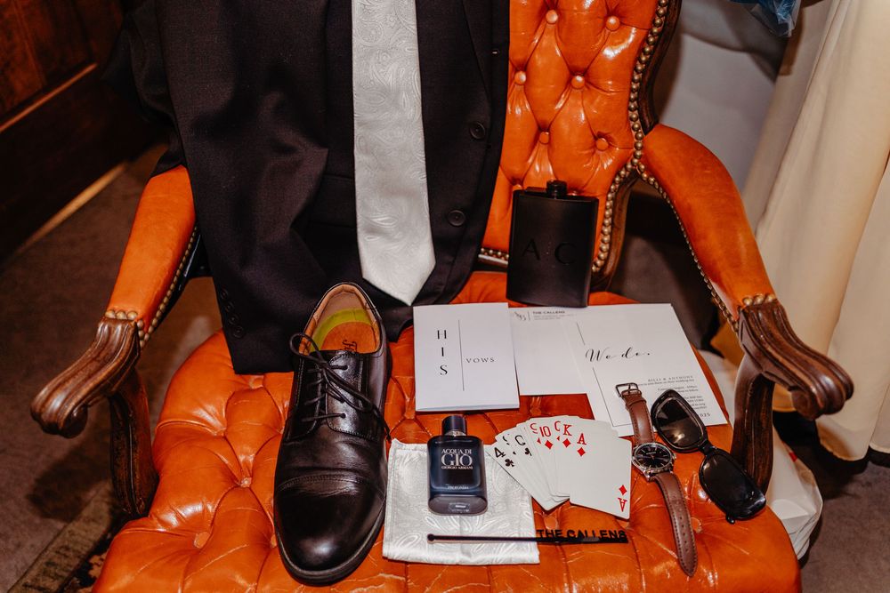 Black dress shoes, cologne, playing cards, and accessories arranged on an orange tufted leather chair for a groom.