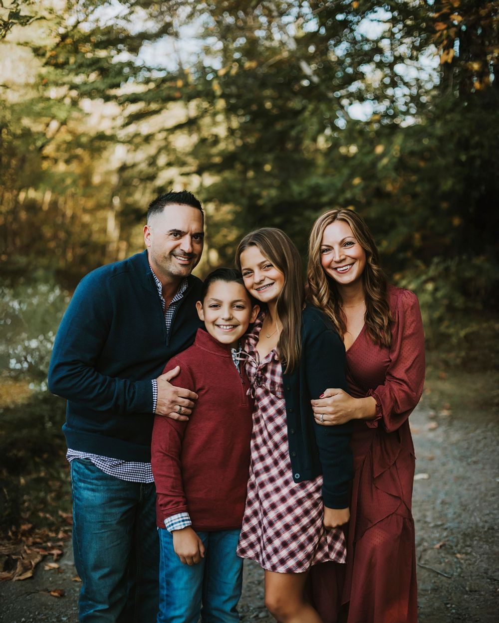 Family of four smiling outdoors in fall, wearing coordinated burgundy and navy outfits, surrounded by autumn foliage.