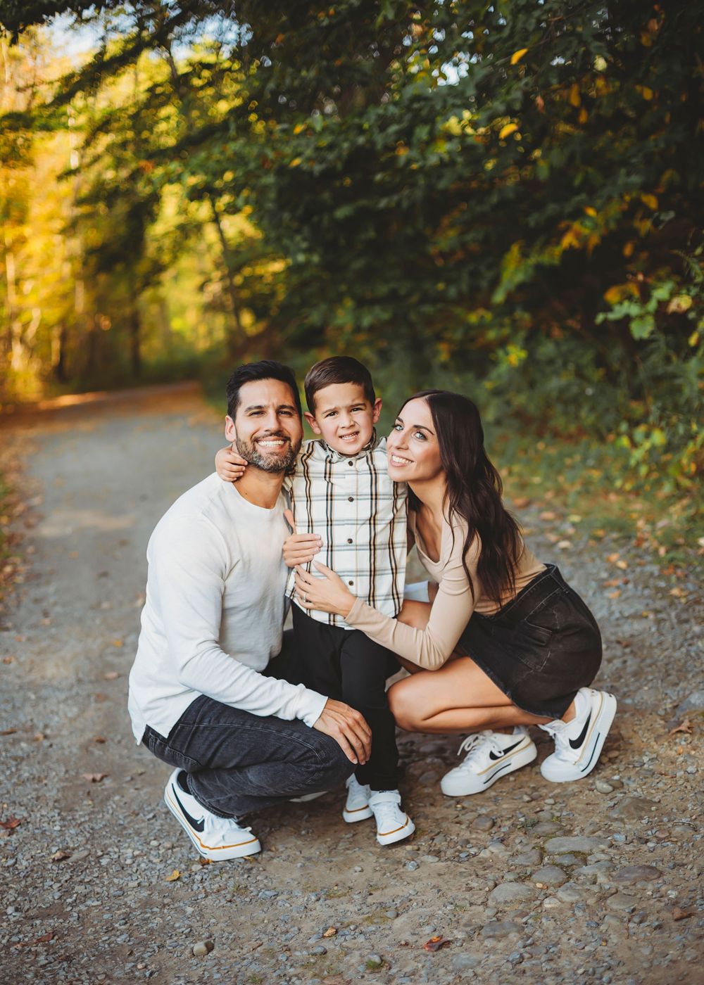 A happy family of three poses on a tree-lined path in autumn, parents hugging their young son outdoors.