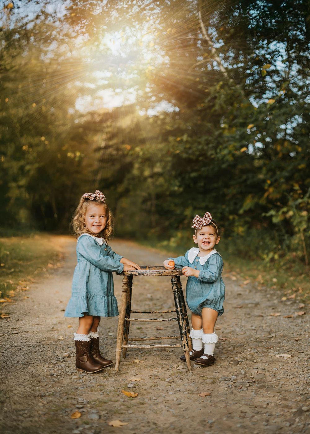 Two smiling toddler girls in matching blue dresses and hair bows pose outdoors on a sunlit dirt path.
