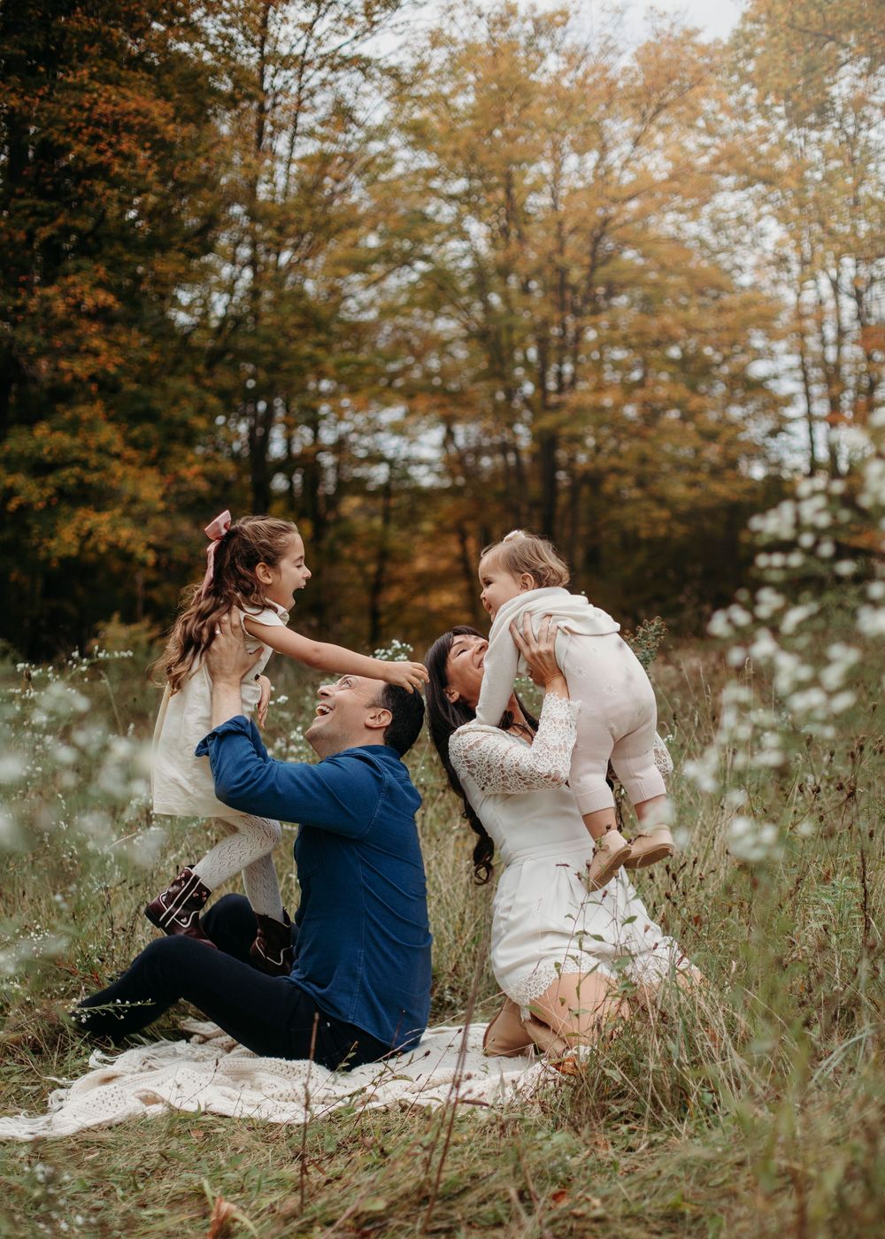 Family of four playing in autumn meadow with colorful fall trees, parents sitting on blanket with two young daughters.