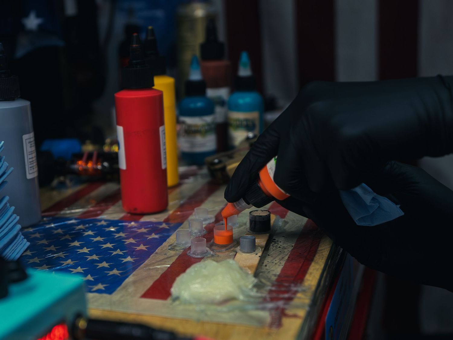 Karl Cooper's gloved hand pours orange ink into a cap on a flag-draped station at Sacred Art Tattoo in Honolulu, Hawaii, ink bottles behind.