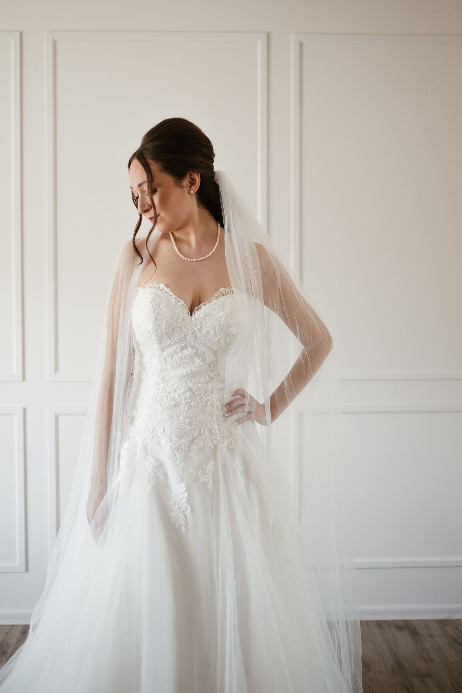 Bride in white lace sweetheart wedding gown with tulle veil and pearl necklace posing against white wall.