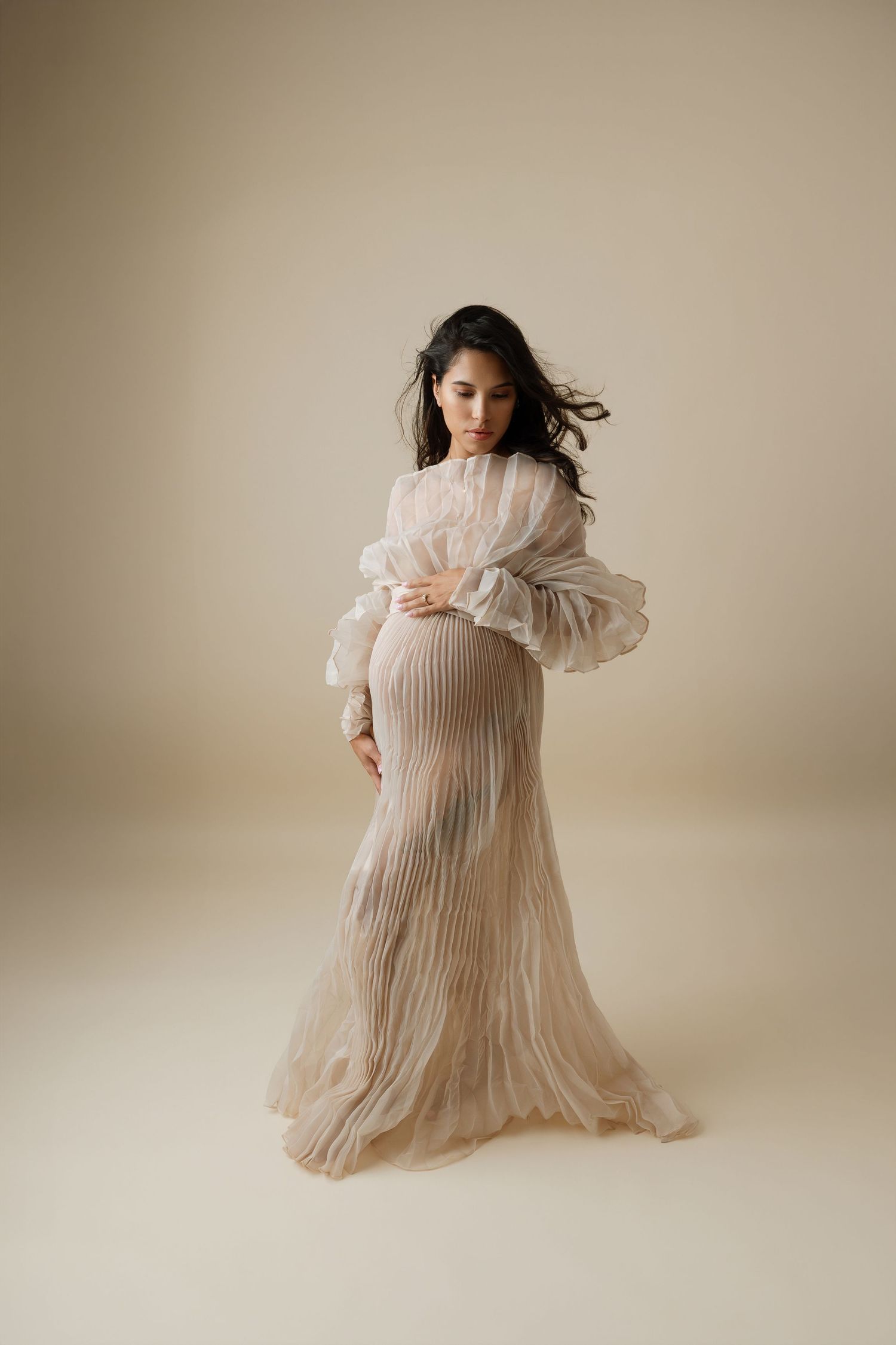 Pregnant woman in sheer pleated beige gown with ruffled sleeves cradling baby bump in elegant maternity studio portrait.