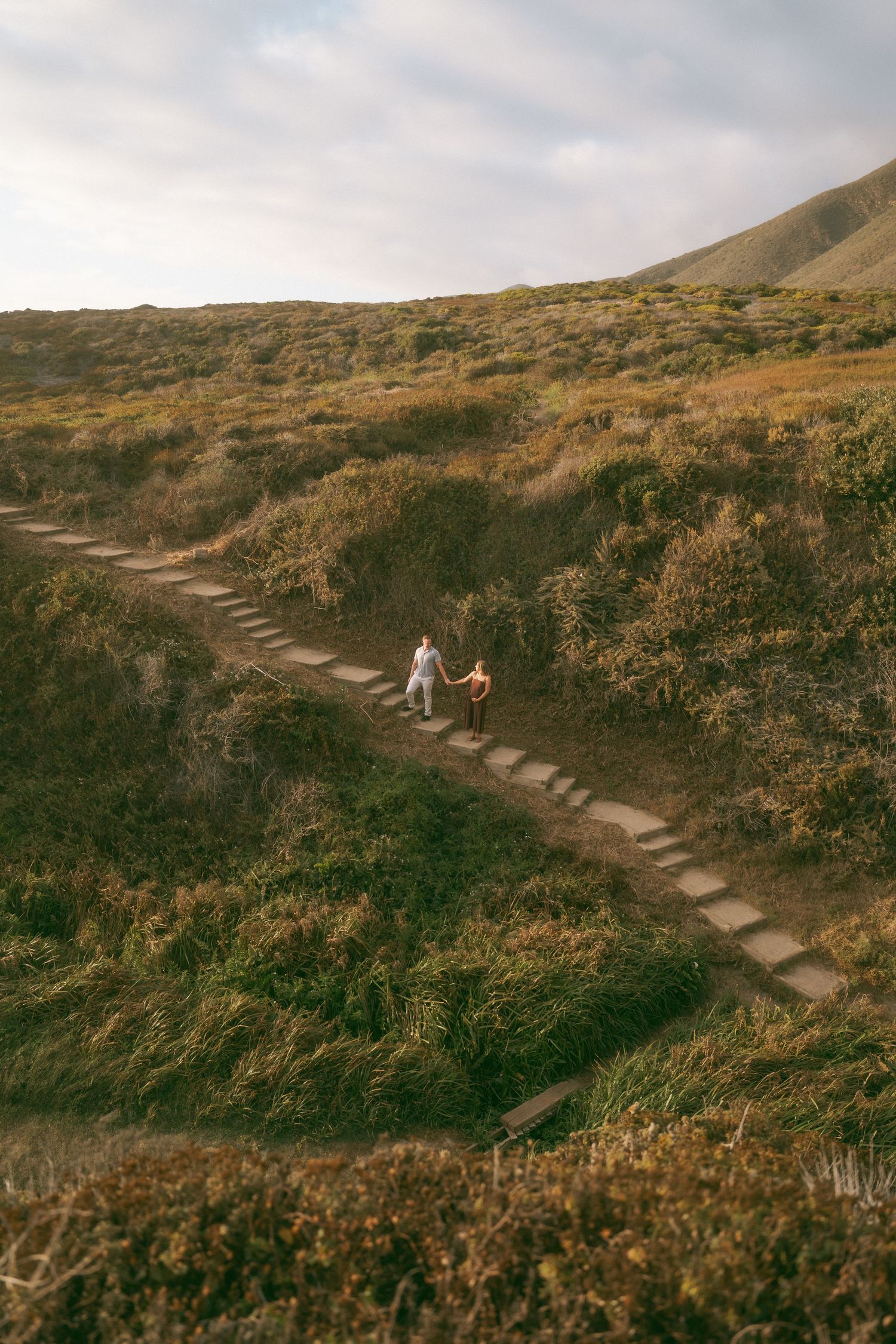 Two hikers walk along a winding stone path through lush green hillside terrain under a cloudy sky.