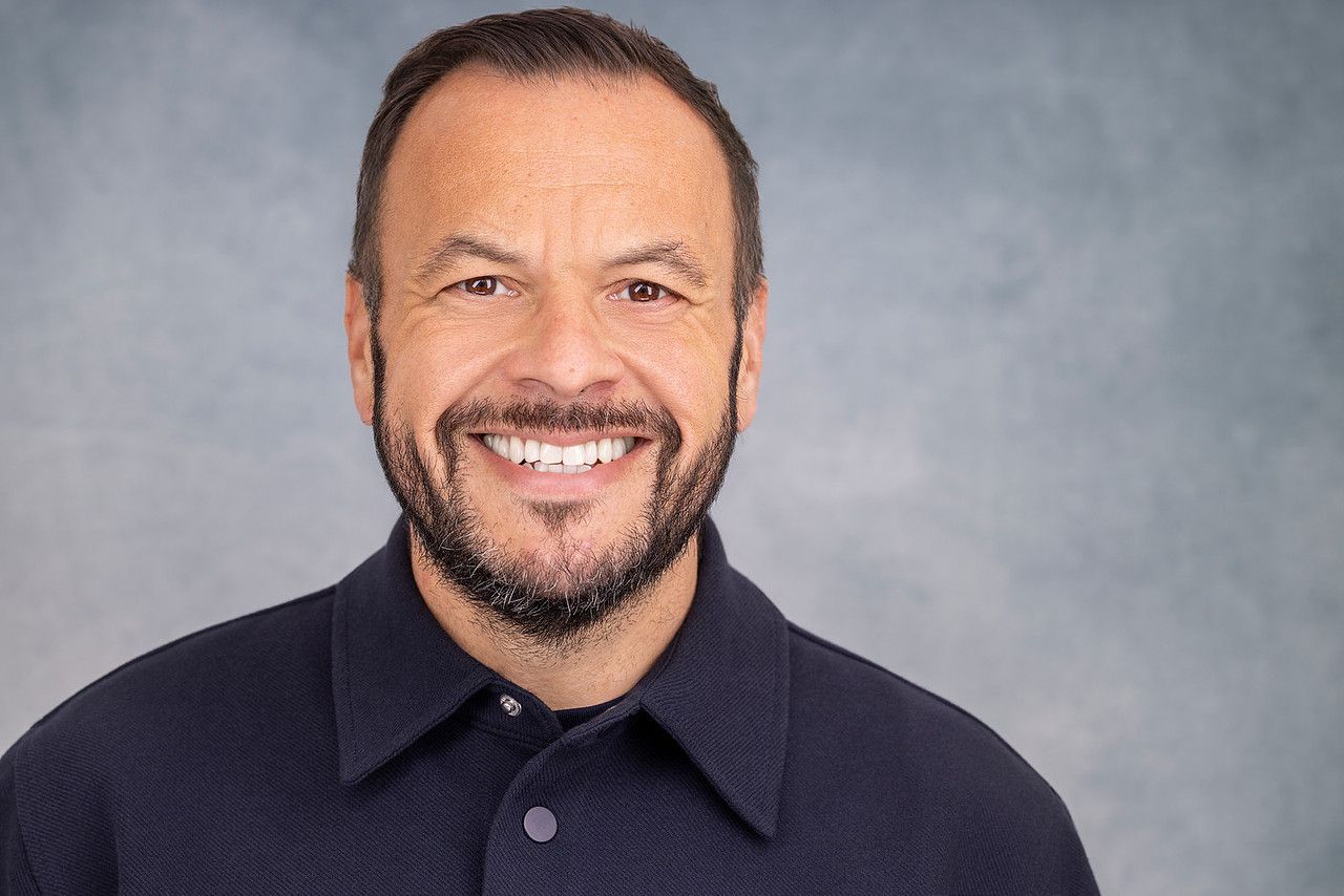 Professional headshot of smiling middle-aged man with beard wearing dark shirt.