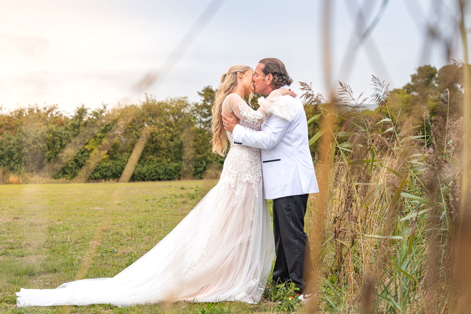 A bride and groom share a romantic kiss in a sunlit field, with tall grass and greenery in the background.Yorkshire, Sheffield, Lincolnshire