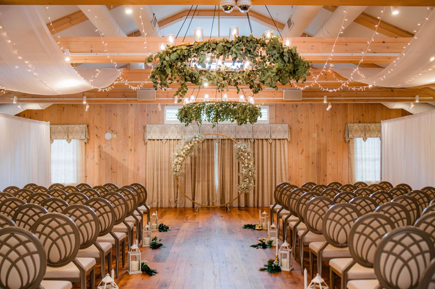 Elegant barn wedding ceremony venue with wooden walls, floral chandelier, fairy lights, and rows of decorated chairs lining the aisle.