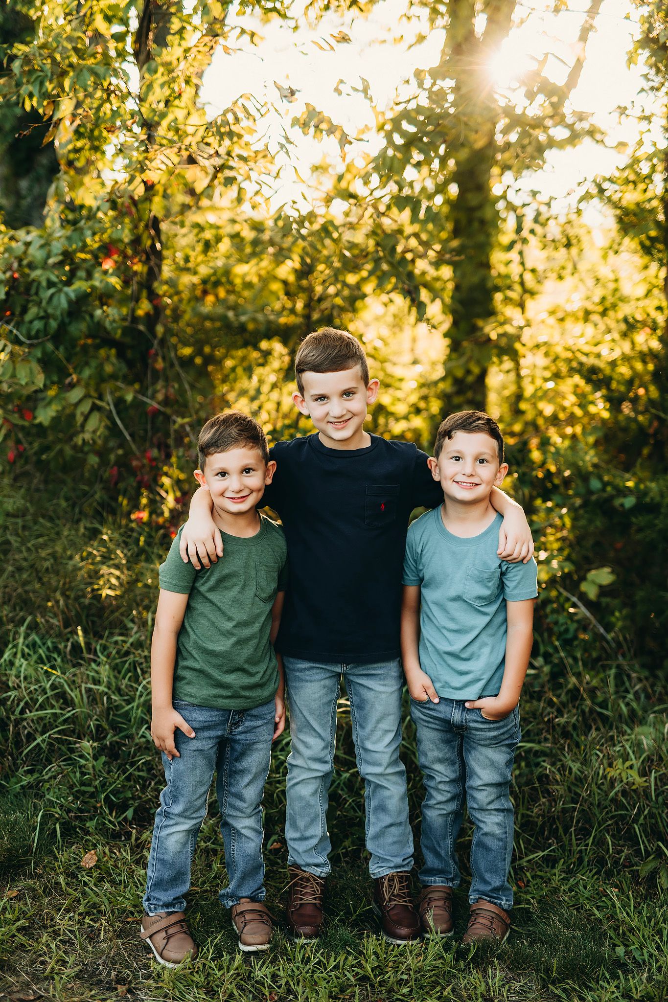 Three smiling brothers in jeans and t-shirts pose together outdoors in a sunlit forest setting.