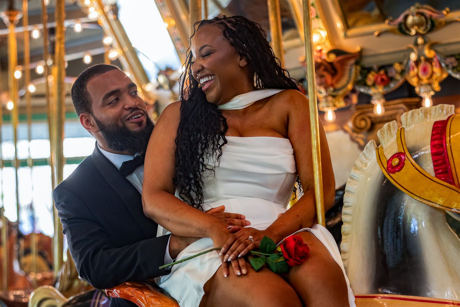 Couple in wedding attire laughing together on a colorful carousel, man in suit and woman in white dress holding a red rose.