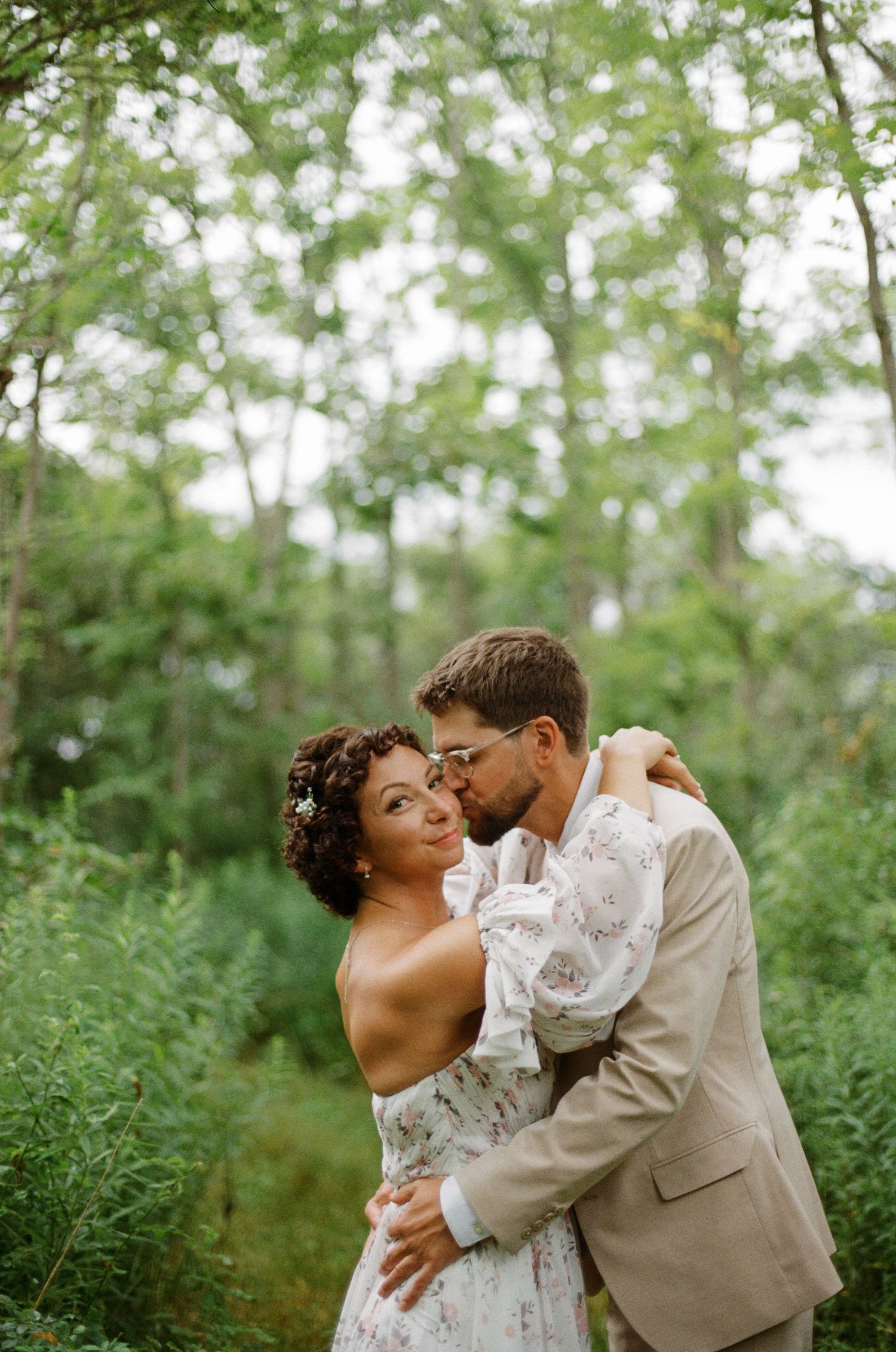 A couple shares a romantic embrace in a lush green forest, the groom kissing the bride's cheek as she smiles.